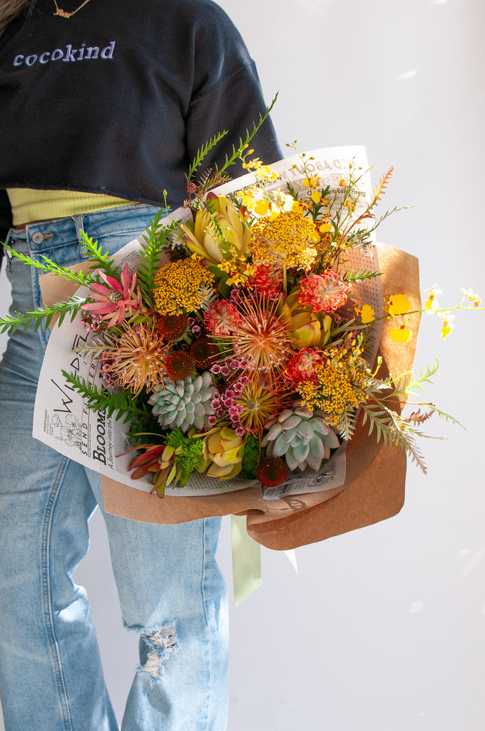 A woman holds a bouquet wrapped in floral newspaper and brown kraft paper in front of a white backdrop. It's filled with yellow, red, and orange flowers, including pincushion protea, grevillea, succulents, leucantha, oncidium orchid, and thistle. The bouquet has a green bow.