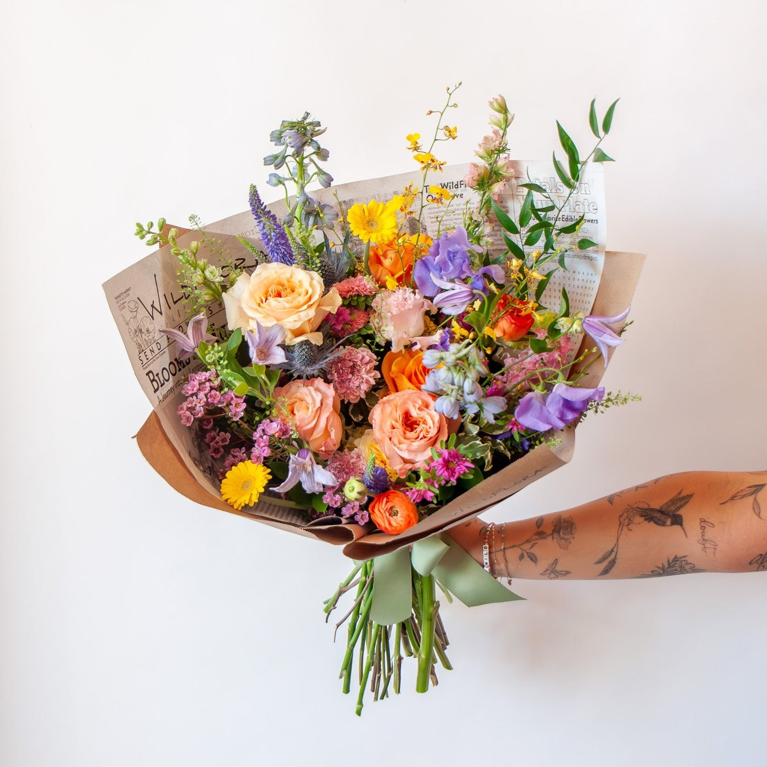 A tattooed arm holds a bouquet wrapped in floral newspaper and brown kraft paper in front of a white backdrop. It's filled with pink, orange, yellow, and purple flowers, including piccolini daisies, roses, veronica, sweet pea, clematis, delphinium, oncidium orchid, and lisianthus.