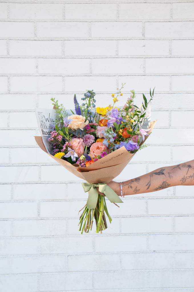 A tattooed arm holds a bouquet wrapped in floral newspaper and brown kraft paper in front of a white brick wall. It's filled with pink, orange, yellow, and purple flowers, including piccolini daisies, roses, veronica, sweet pea, clematis, delphinium, oncidium orchid, and lisianthus.