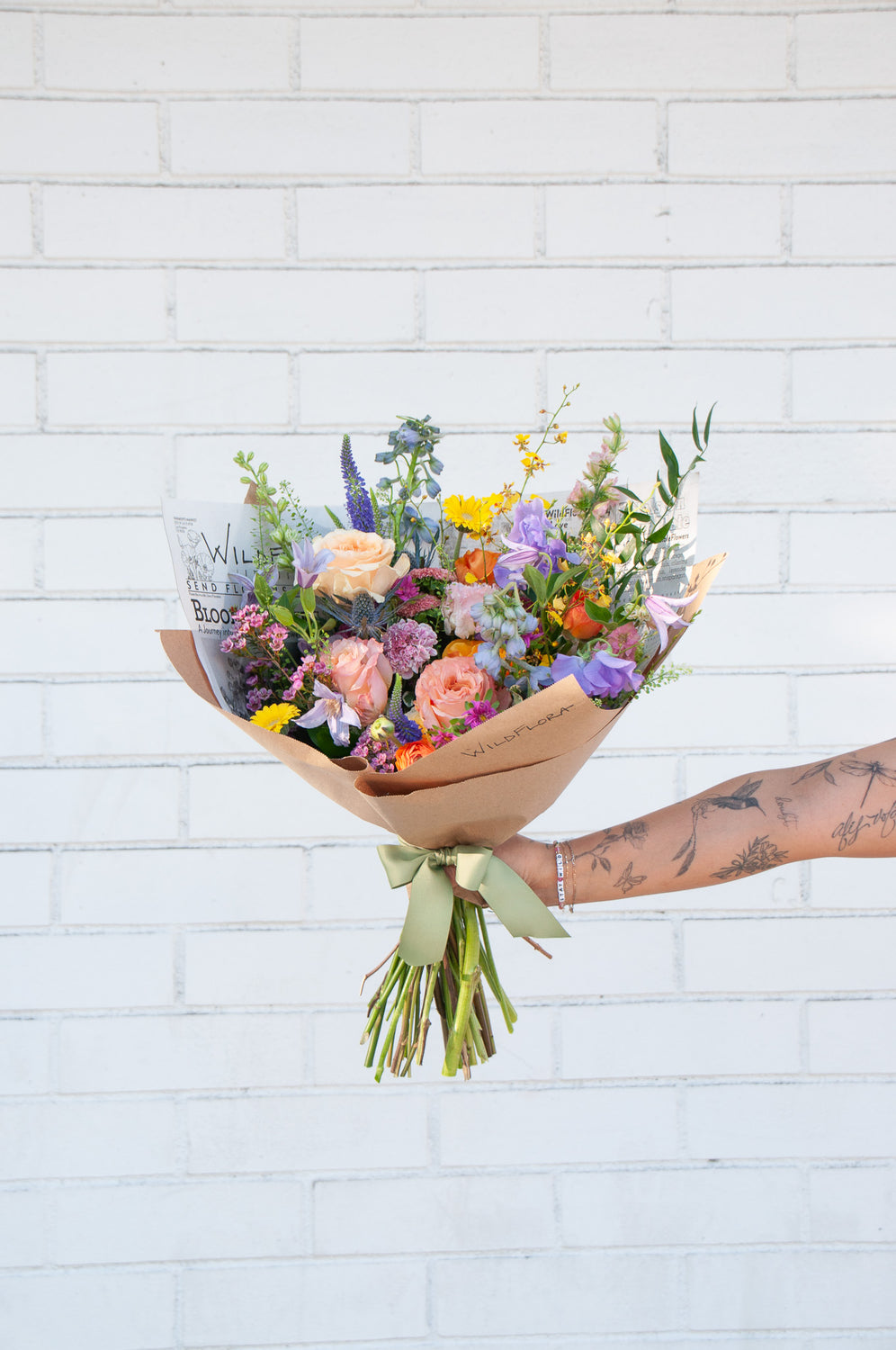 A tattooed arm holds a bouquet wrapped in floral newspaper and brown kraft paper in front of a white brick wall. It's filled with pink, orange, yellow, and purple flowers, including piccolini daisies, roses, veronica, sweet pea, clematis, delphinium, oncidium orchid, and lisianthus.