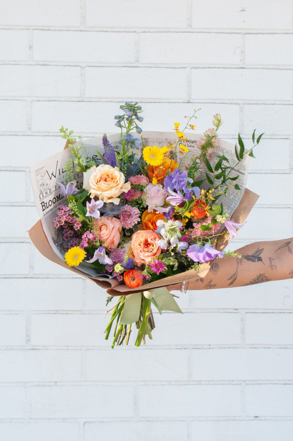 A tattooed arm holds a bouquet wrapped in floral newspaper and brown kraft paper in front of a white brick wall. It's filled with pink, orange, yellow, and purple flowers, including piccolini daisies, roses, veronica, sweet pea, clematis, delphinium, oncidium orchid, and lisianthus.