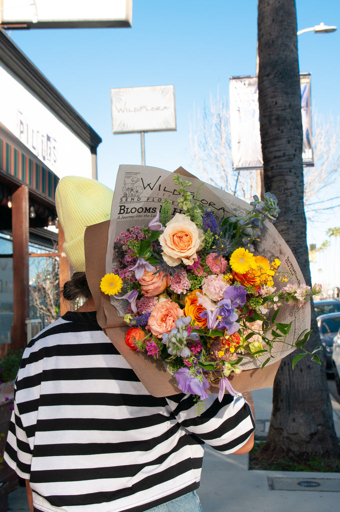 a woman in a striped shirt on ventura blvd holds a bouquet wrapped in floral newspaper and brown kraft paper over her shoulder toward the camera on a sunny day. It's filled with pink, orange, yellow, and purple flowers, including piccolini daisies, roses, veronica, sweet pea, clematis, delphinium, oncidium orchid, and lisianthus.