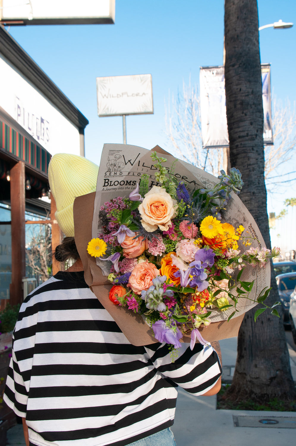 a woman in a striped shirt on ventura blvd holds a bouquet wrapped in floral newspaper and brown kraft paper over her shoulder toward the camera on a sunny day. It's filled with pink, orange, yellow, and purple flowers, including piccolini daisies, roses, veronica, sweet pea, clematis, delphinium, oncidium orchid, and lisianthus.