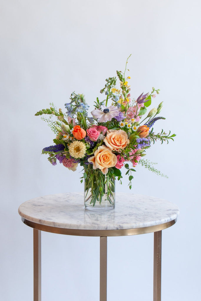 Petite Seasonal WildFlora flower arrangement. The flowers are many colors and include roses, tulips, snapdragon, veronica, anemone, clematis, orchids, and delphinium.