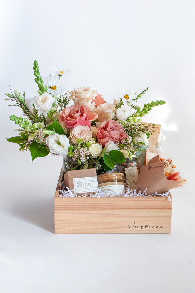 A floral arrangement featuring pink roses, white flowers, and greenery, presented in a wooden gift box with a bath soak, a dried mini bouquet, shower steamers, bath salts, and a small candle.