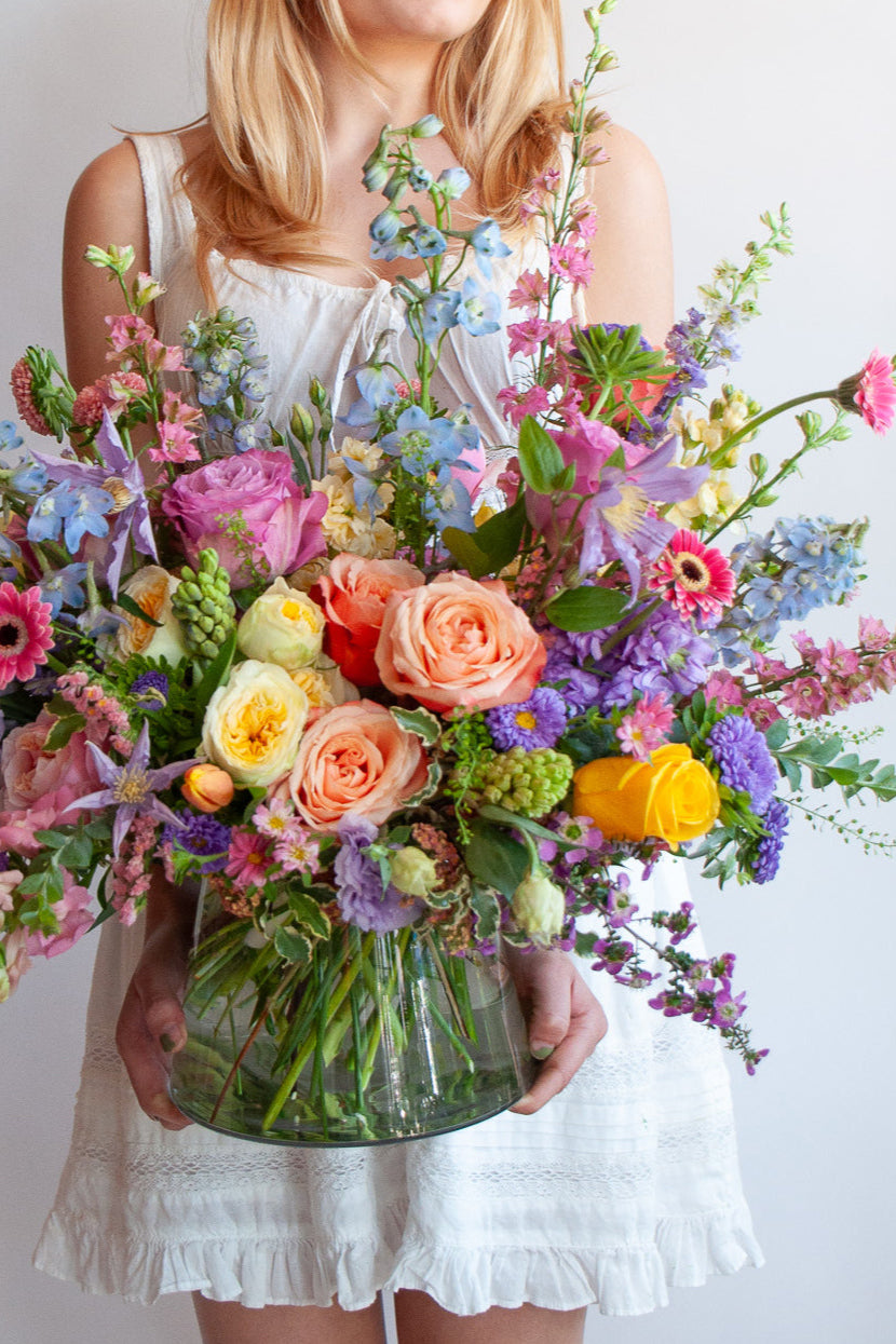 a woman in a white dress holds a glass vase with a large flower arrangement in it. The flowers are many colors and include roses, tulips, daisies, snapdragon, clematis, stock, hyacinth, and delphinium.