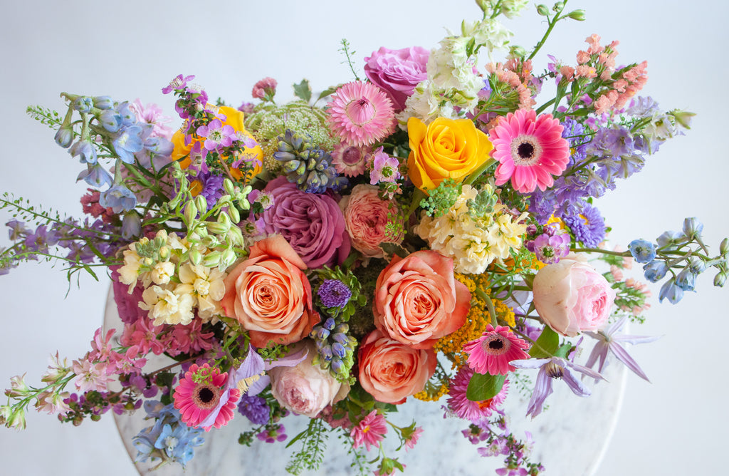 Close up of the blooms in a flower arrangement. The flowers are many colors and include roses, tulips, daisies, snapdragon, clematis, stock, straw flower, and delphinium.