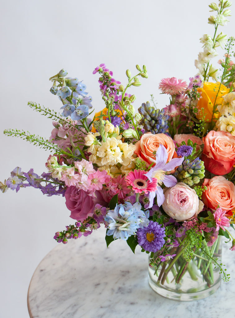 Close up of the blooms in a flower arrangement. The flowers are many colors and include roses, tulips, daisies, snapdragon, clematis, stock, straw flower, and delphinium.