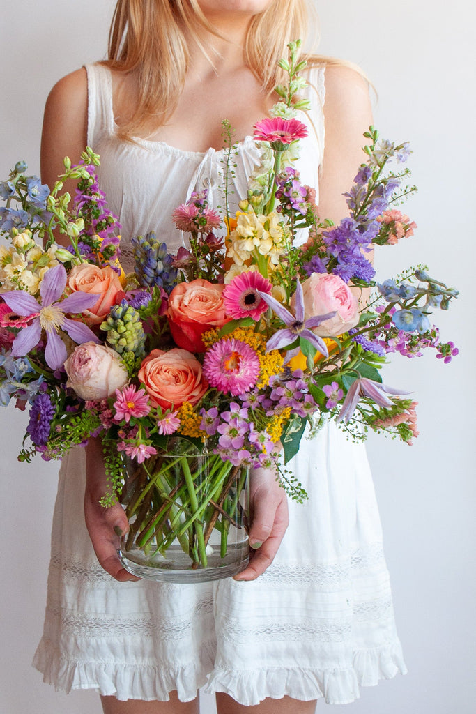 a woman in a white dress holds a glass vase with a medium flower arrangement in it. The flowers are many colors and include roses, tulips, daisies, snapdragon, clematis, stock, straw flower, and delphinium.