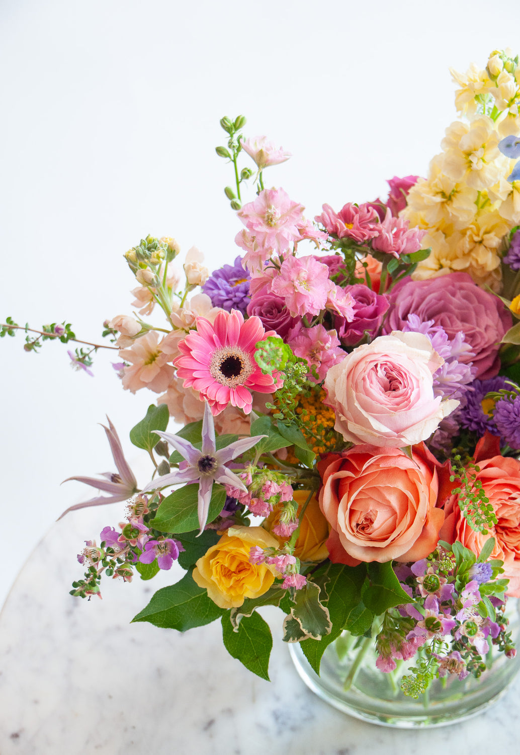 Close up of the blooms in a flower arrangement. The flowers are many colors and include roses, tulips, daisies, snapdragon, clematis, stock, and delphinium.