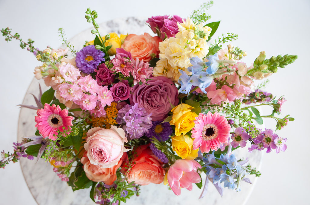 Close up of the blooms in a flower arrangement. The flowers are many colors and include roses, tulips, daisies, snapdragon, clematis, stock, and delphinium.