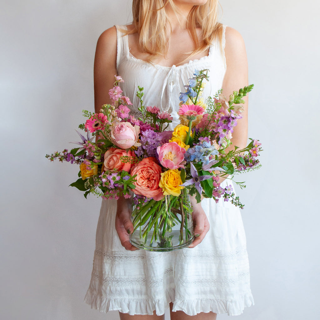 Woman holding a glass vase with a small flower arrangement in front of a white backdrop. The flowers are many colors and include roses, tulips, daisies, snapdragon, clematis, and delphinium.