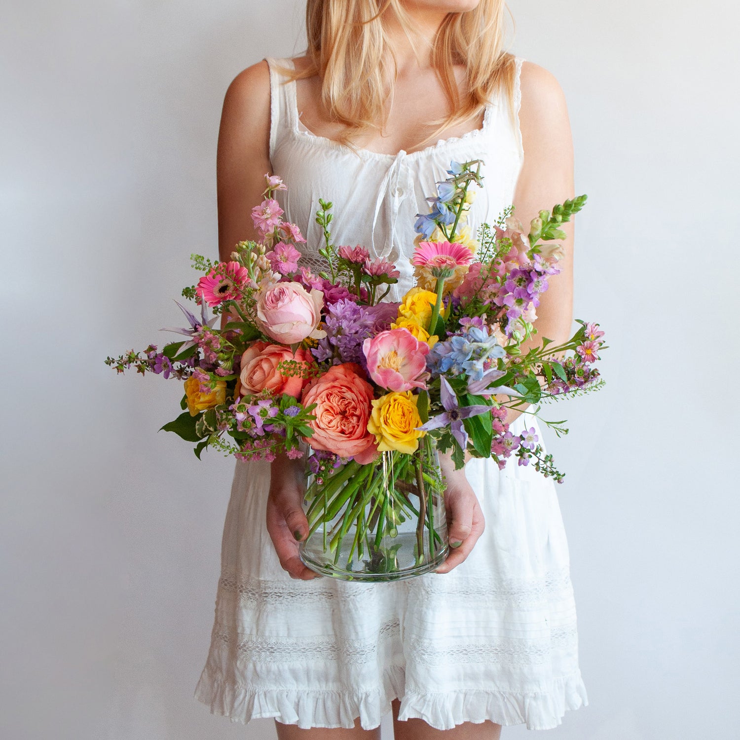Woman holding a glass vase with a small flower arrangement in front of a white backdrop. The flowers are many colors and include roses, tulips, daisies, snapdragon, clematis, and delphinium.