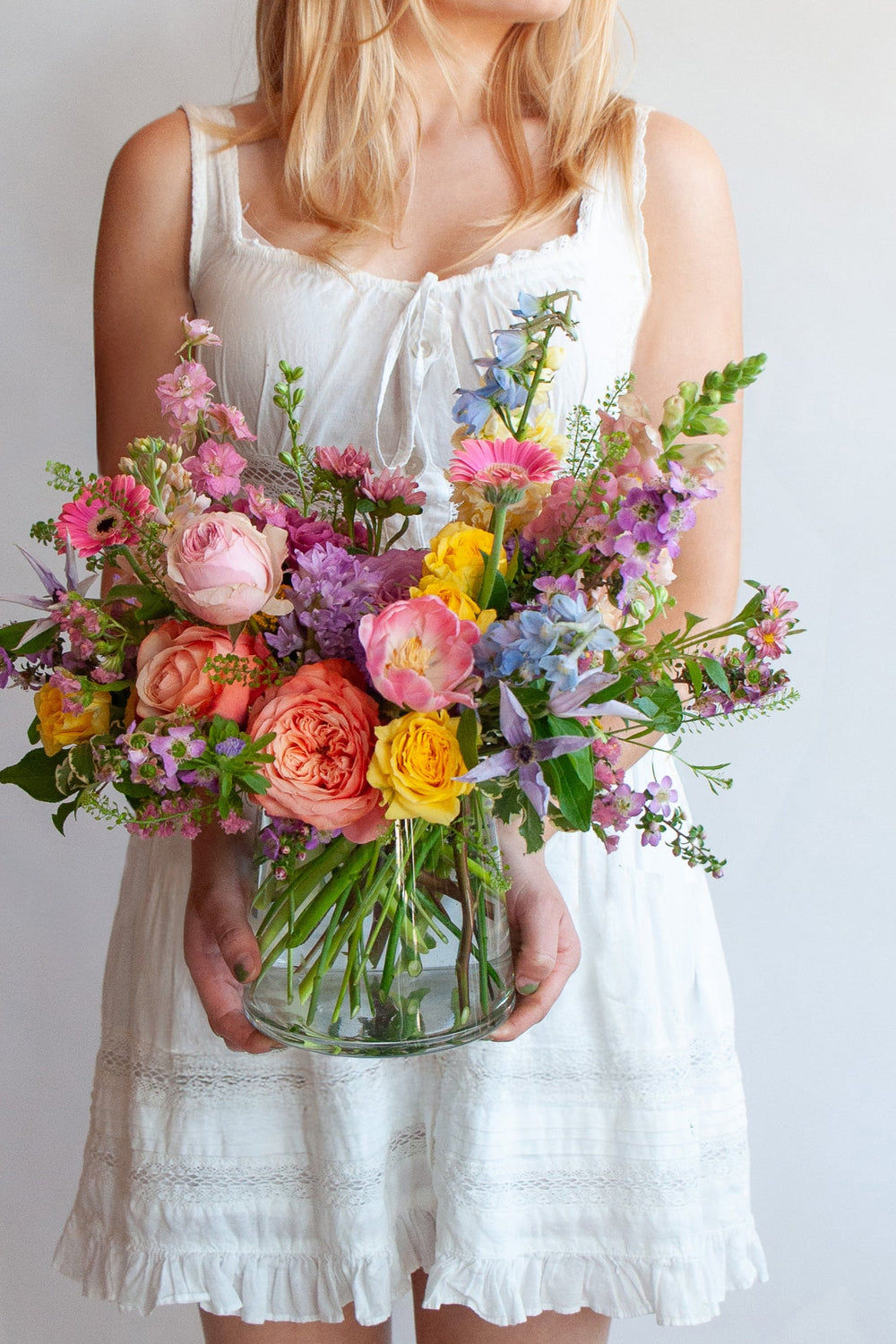 Woman holding a glass vase with a small flower arrangement in front of a white backdrop. The flowers are many colors and include roses, tulips, daisies, snapdragon, clematis, and delphinium.