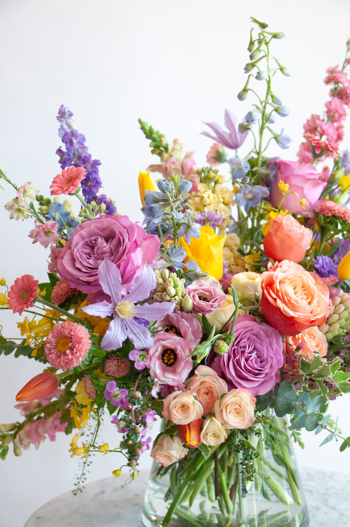 Close up of the blooms in a flower arrangement. The flowers are many colors and include roses, tulips, daisies, snapdragon, clematis, stock, hyacinth, orchids, and delphinium.