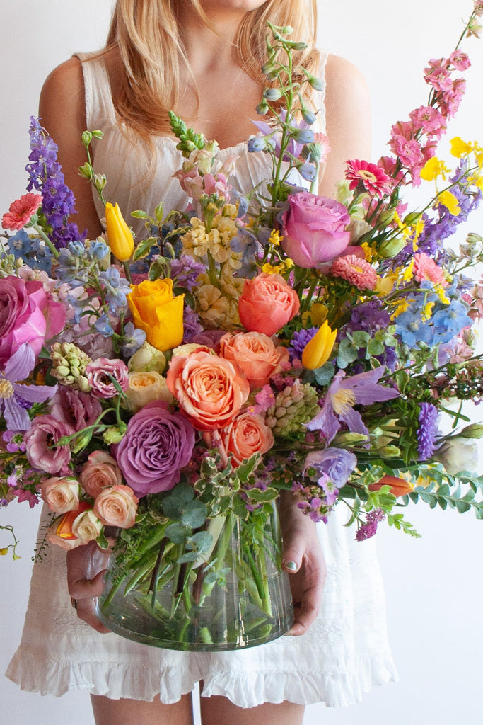 a woman in a white dress holds a glass vase with an extra large flower arrangement in it. The flowers are many colors and include roses, tulips, daisies, snapdragon, clematis, stock, hyacinth, orchids, and delphinium.