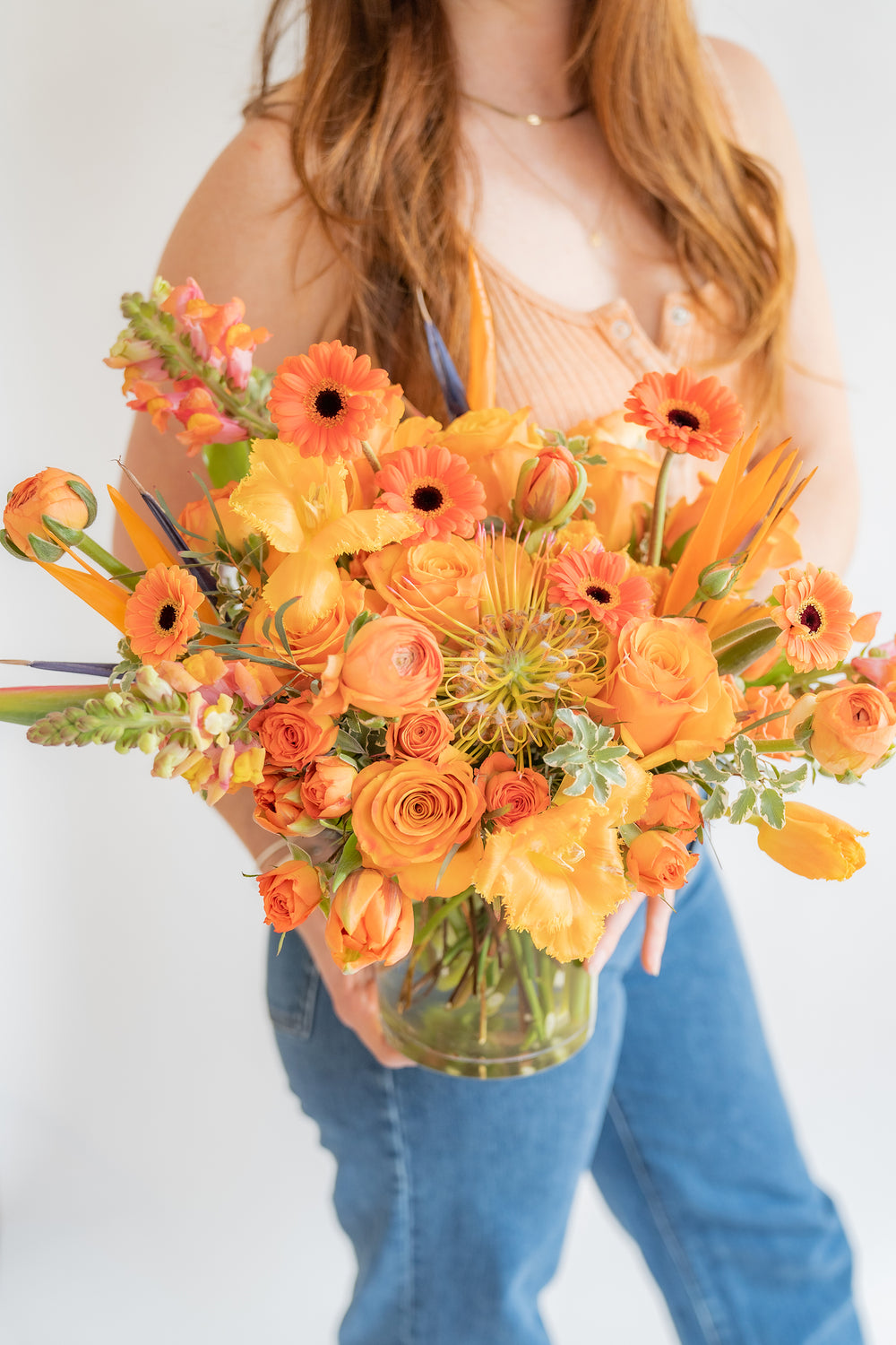 a woman in orange holding A flower arrangement in a glass container in front of a white backdrop. It's filled with orange and tangerine-colored flowers, including rose, snapdragon, piccolini daisy, bird of paradise, tulips, and pincushion protea.