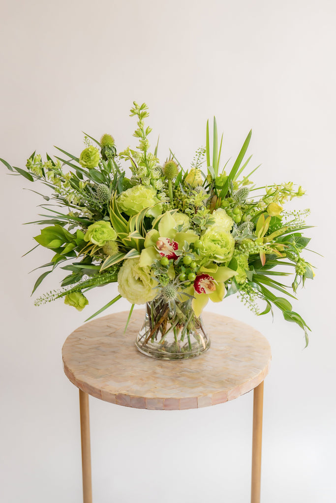 A flower arrangement in a glass container in front of a white backdrop on a small table. It's filled with green and chartreuse flowers and foliage, including rose, lisianthus, eucalyptus, and cymbidium orchid.
