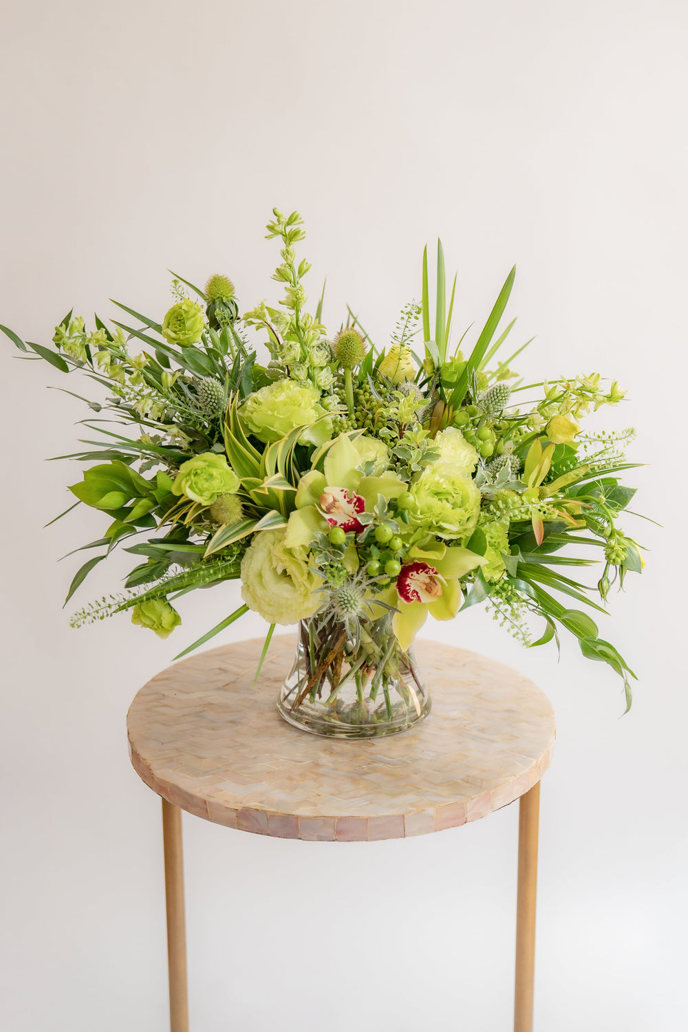 A flower arrangement in a glass container in front of a white backdrop on a small table. It's filled with green and chartreuse flowers and foliage, including rose, lisianthus, eucalyptus, and cymbidium orchid.