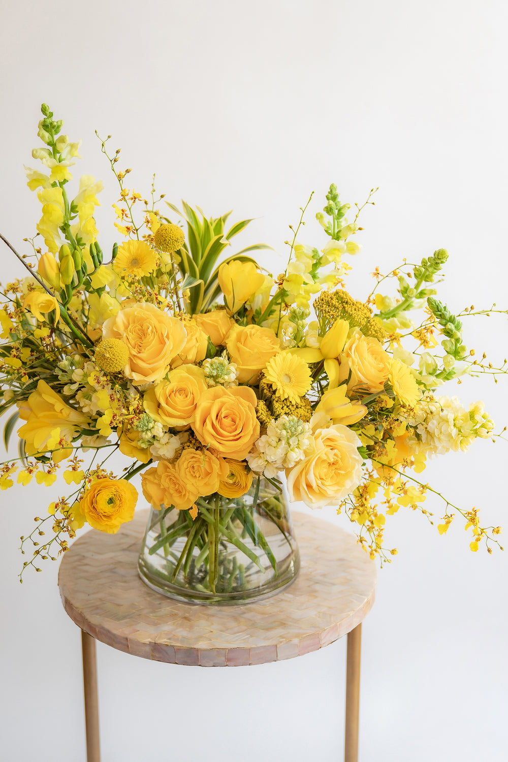 A flower arrangement in a glass container in front of a white backdrop on a small table. It's filled with yellow and gold flowers, including rose, snapdragon, billy balls, spray rose, yarrow, stock, daffodils, and oncidium orchid.