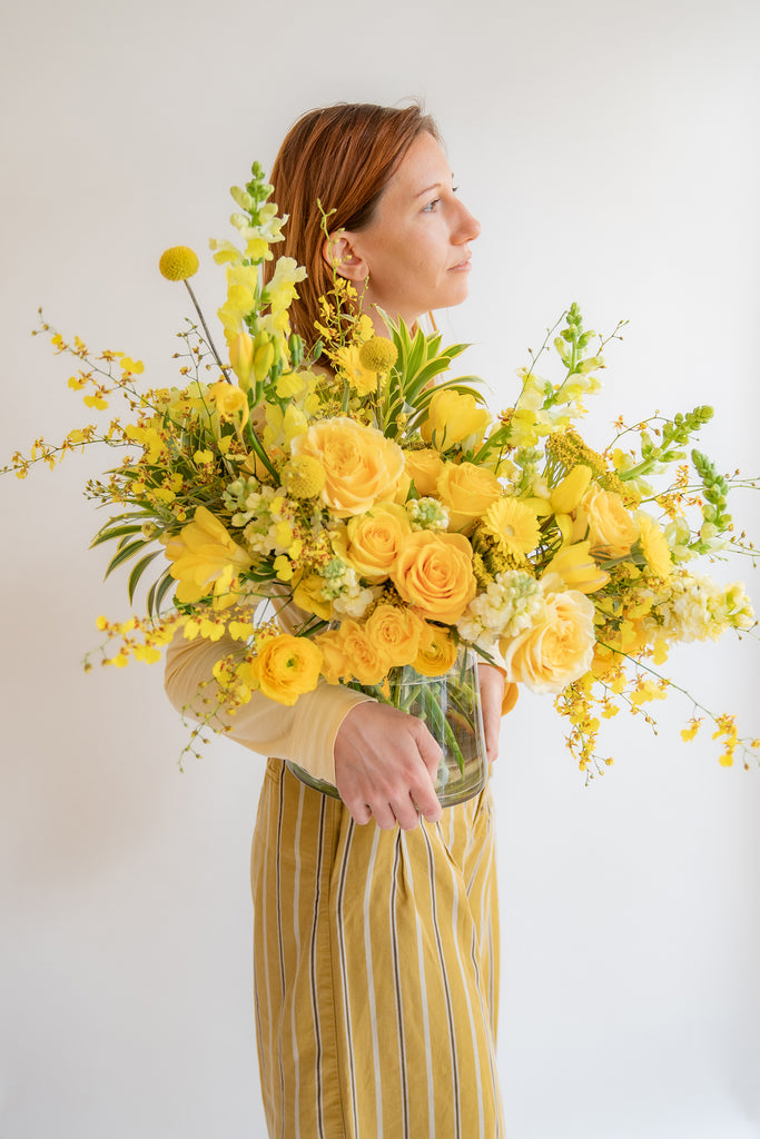 A woman holding a monochromatic flower arrangement in a glass container in front of a white backdrop. It's filled with yellow and gold flowers, including rose, snapdragon, billy balls, spray rose, yarrow, stock, daffodils, and oncidium orchid.