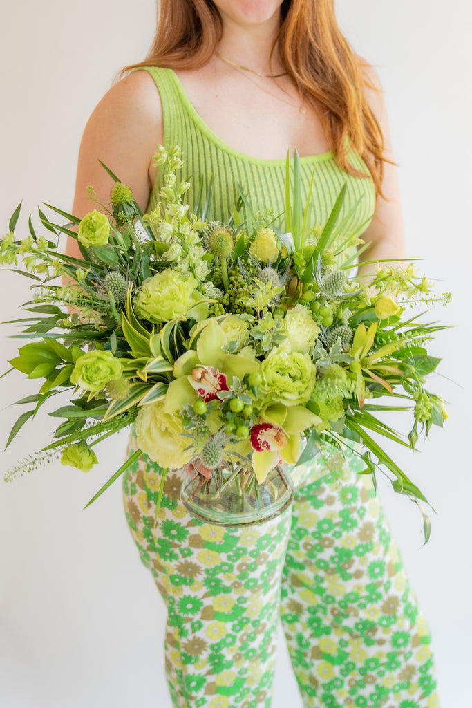 a woman in all green holding A flower arrangement in a glass container in front of a white backdrop. It's filled with green and chartreuse flowers and foliage, including rose, lisianthus, eucalyptus, and cymbidium orchid.