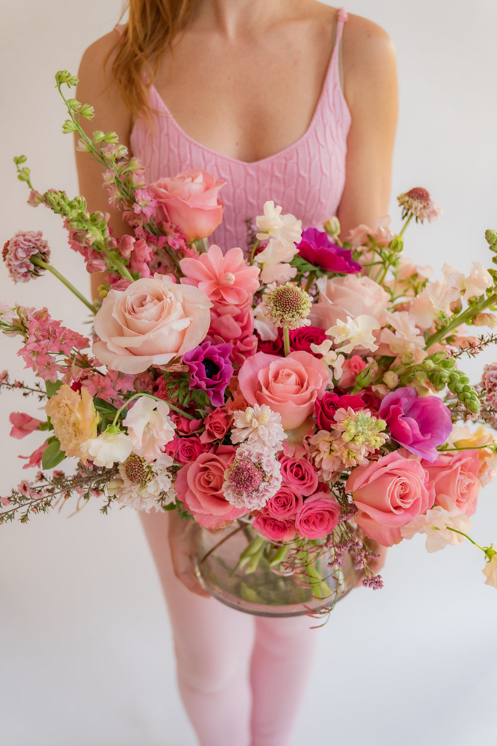 A woman in pink holding A flower arrangement in a glass container in front of a white backdrop. It's filled with pink and magenta flowers, including rose, scabiosa, sweet pea, snapdragon, and delphinium.