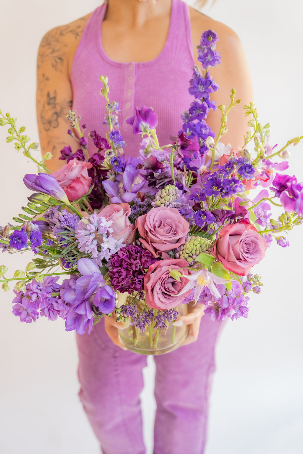 a woman in all purple holds A flower arrangement in a glass container in front of a white backdrop. It's filled with lavender and purple flowers, including rose, stock, delphinium, and statice, hyacinth, and clematis.