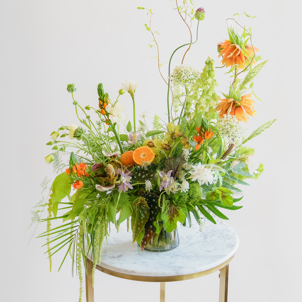 A large flower arrangement in a glass vase in front of a white backdrop on a marble table. It's filled with orange, white, purple, but mostly green and and chartreuse-colored flowers and foliage, including fritillaria, star of bethlehem, orange slices, alium, pitcher plant, anemone, fern, philodendron, polka dot begonia, rose, clematis, bells of ireland, heuchera, and amaranthus. The vibes are very Hobbiton.