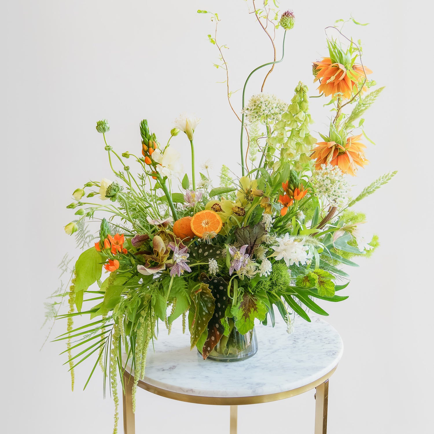A large flower arrangement in a glass vase in front of a white backdrop on a marble table. It's filled with orange, white, purple, but mostly green and and chartreuse-colored flowers and foliage, including fritillaria, star of bethlehem, orange slices, alium, pitcher plant, anemone, fern, philodendron, polka dot begonia, rose, clematis, bells of ireland, heuchera, and amaranthus. The vibes are very Hobbiton.