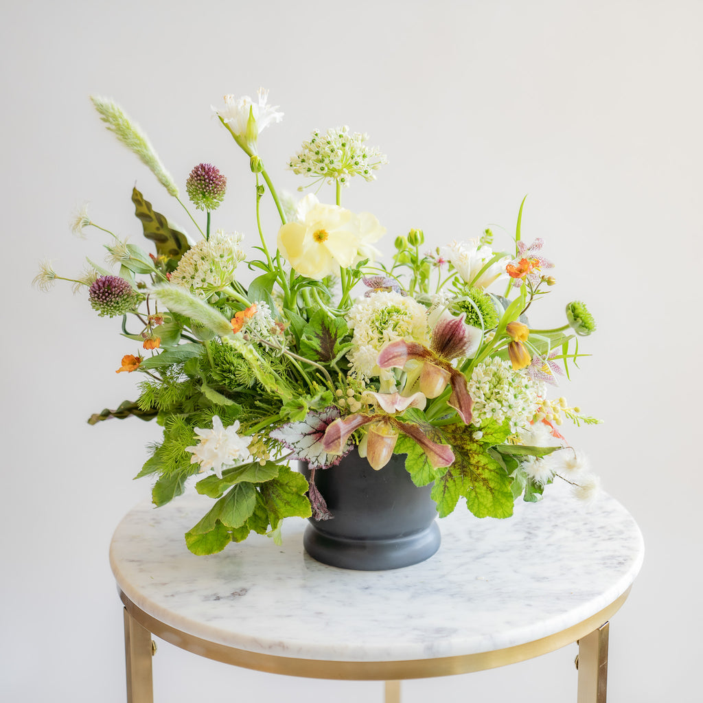 A flower arrangement in a matte black vase in front of a white backdrop on a marble table. It's filled with orange, white, purple, but mostly green and and chartreuse-colored flowers and foliage, including sword fern, fritillaria, alium, pitcher plant, anemone, ornamental grass, calathea peacock plant, clematis, and heuchera. The vibes are very Hobbiton.