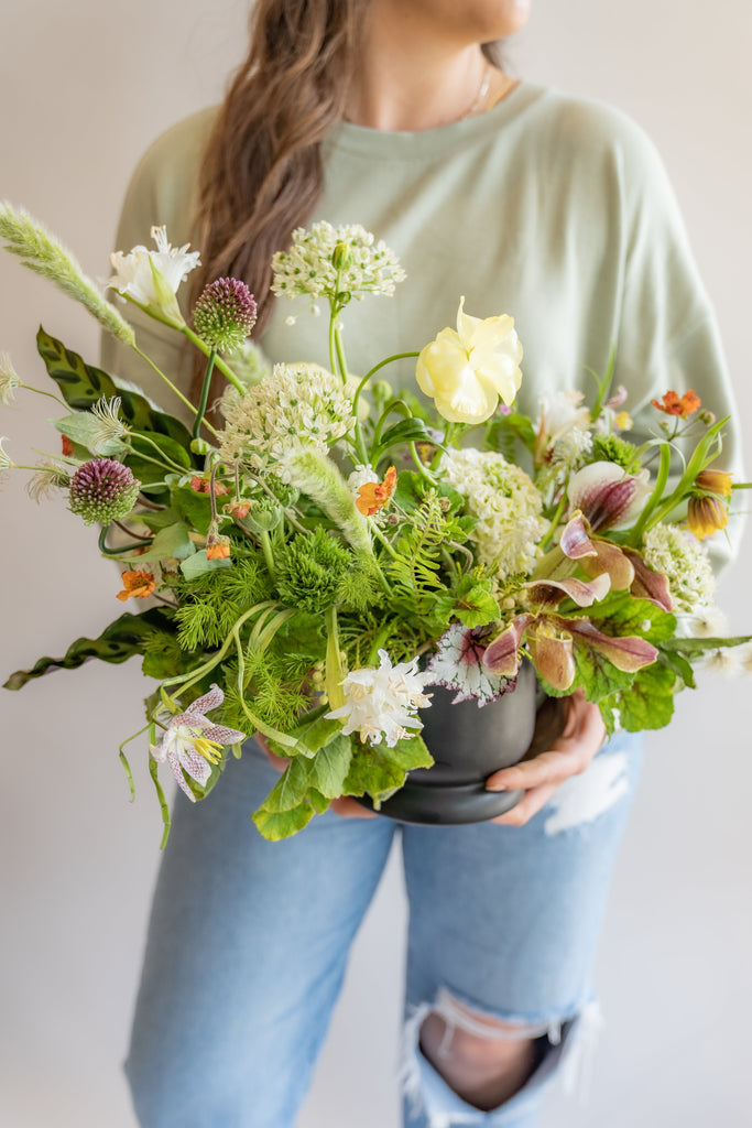 Woman holding A flower arrangement in a matte black vase in front of a white backdrop. It's filled with orange, white, purple, but mostly green and and chartreuse-colored flowers and foliage, including sword fern, fritillaria, alium, pitcher plant, anemone, ornamental grass, calathea peacock plant, clematis, and heuchera. The vibes are very Hobbiton.