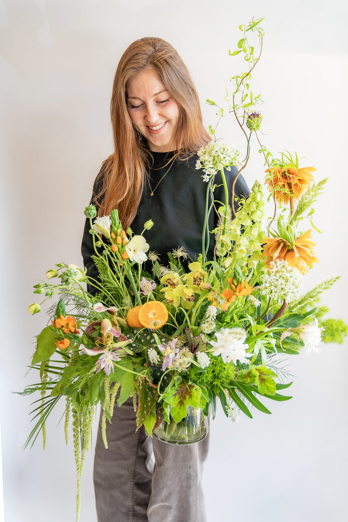woman smiling and holding A large flower arrangement in a glass vase in front of a white backdrop. It's filled with orange, white, purple, but mostly green and and chartreuse-colored flowers and foliage, including fritillaria, star of bethlehem, orange slices, alium, pitcher plant, anemone, fern, philodendron, polka dot begonia, rose, clematis, bells of ireland, heuchera, and amaranthus. The vibes are very Hobbiton.