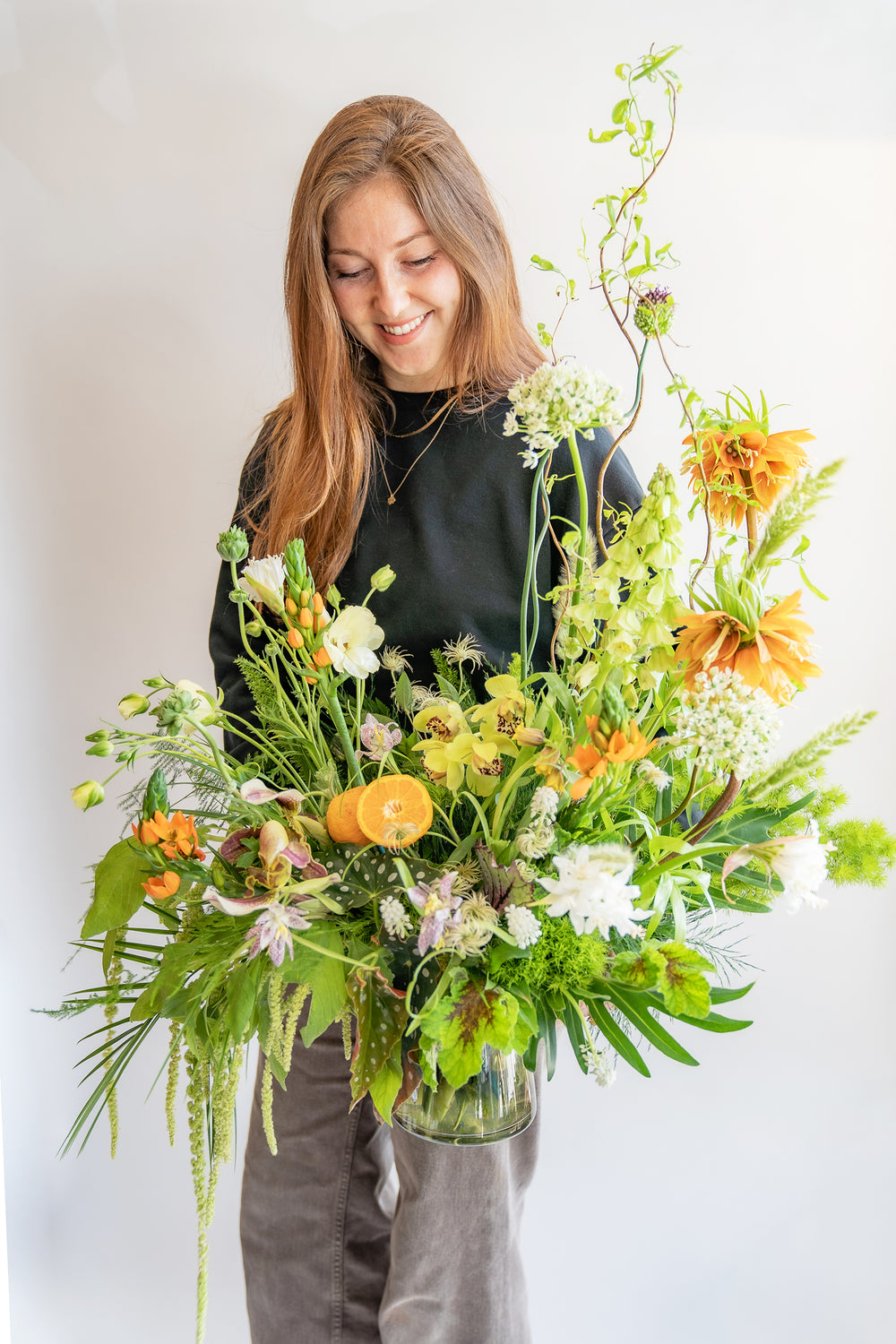 woman smiling and holding A large flower arrangement in a glass vase in front of a white backdrop. It's filled with orange, white, purple, but mostly green and and chartreuse-colored flowers and foliage, including fritillaria, star of bethlehem, orange slices, alium, pitcher plant, anemone, fern, philodendron, polka dot begonia, rose, clematis, bells of ireland, heuchera, and amaranthus. The vibes are very Hobbiton.