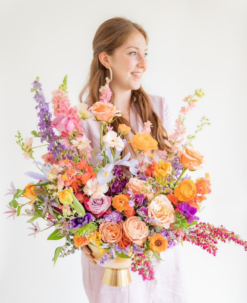 A woman in a lavender dress holds a large flower arrangement in a gold urn in front of a white backdrop. It's filled with pink, magenta, purple, peach, orange, and violet-colored flowers, including rose, ranunculus, clematis, delphinium, anemone, tulip, and snapdragon.