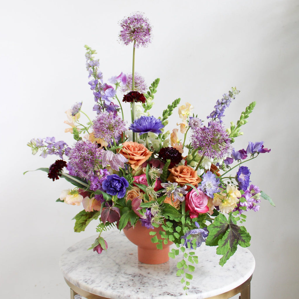 A large flower arrangement in a terracotta container on a marble table with green, purple, indigo, pink, orange, dark violet, and cream-colored flowers, including rose, ranunculus, snapdragon, sweet pea, maidenhair fern, scabiosa, stock, fritillaria, and allium. The backdrop is white.