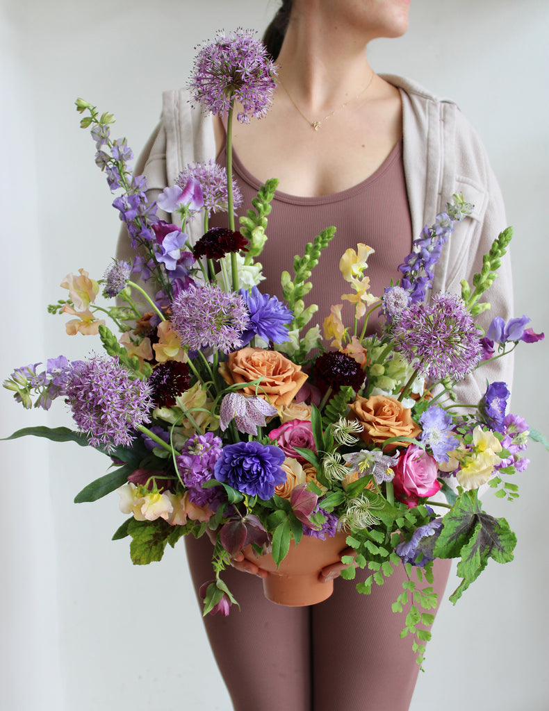 A woman in a brown bodysuit holds A large flower arrangement in a terracotta container with green, purple, indigo, pink, orange, dark violet, and cream-colored flowers, including rose, ranunculus, snapdragon, sweet pea, maidenhair fern, scabiosa, stock, fritillaria, and allium. The backdrop is white.