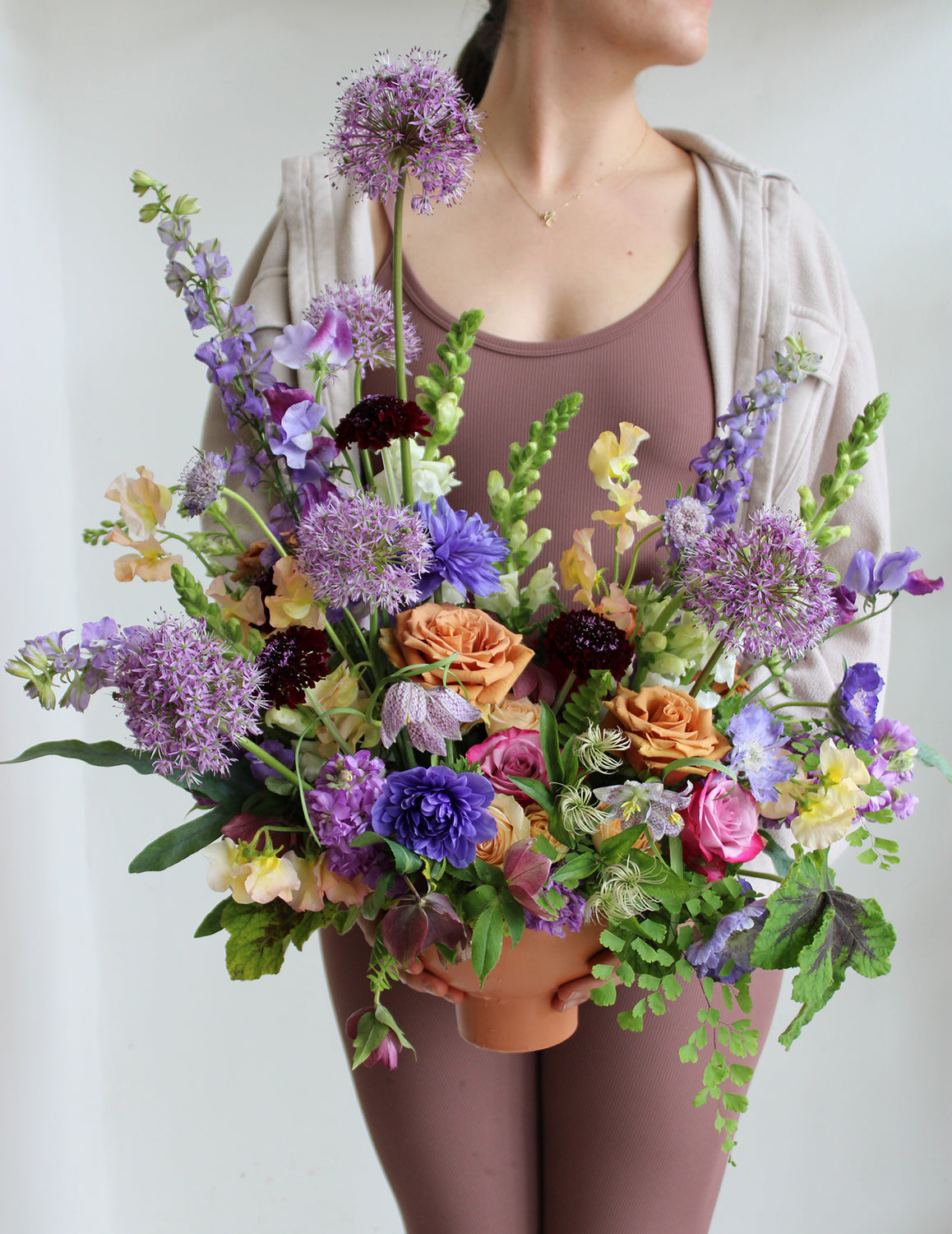 A woman in a brown bodysuit holds A large flower arrangement in a terracotta container with green, purple, indigo, pink, orange, dark violet, and cream-colored flowers, including rose, ranunculus, snapdragon, sweet pea, maidenhair fern, scabiosa, stock, fritillaria, and allium. The backdrop is white.
