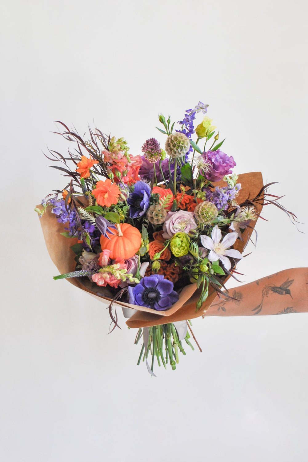 Bouquet of purple green and orange flowers, with a tiny pumpkin, held by a hand with tattoos on a white background