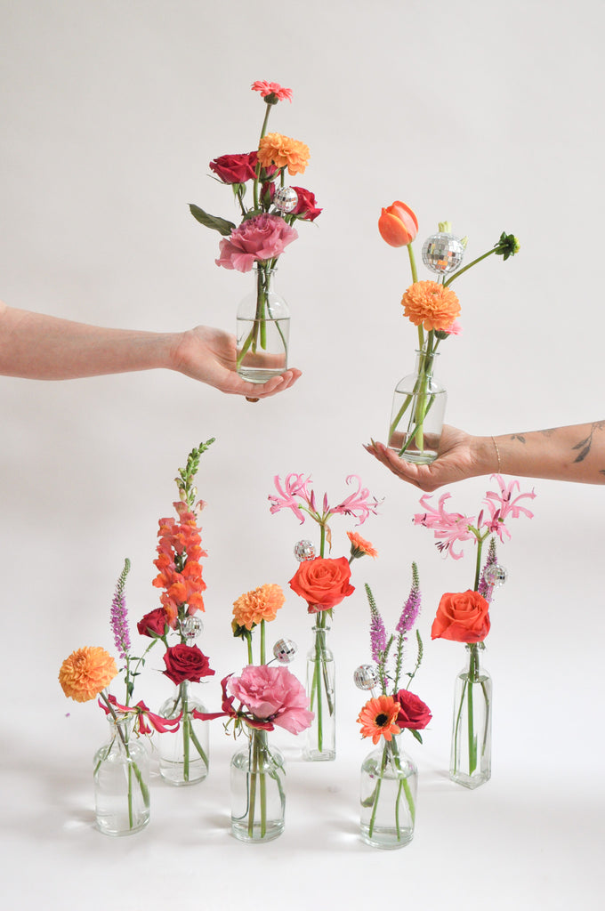 two hands hold clear glass bud vases with tiny round, reflective mirror ball disco balls as well. Below on the white backdrop, sit more of these vases. They're filled with pink, orange, tangerine, and magenta-colored flowers, including rose, dahlia, piccolini daisy, tulip, and veronica and lily.
