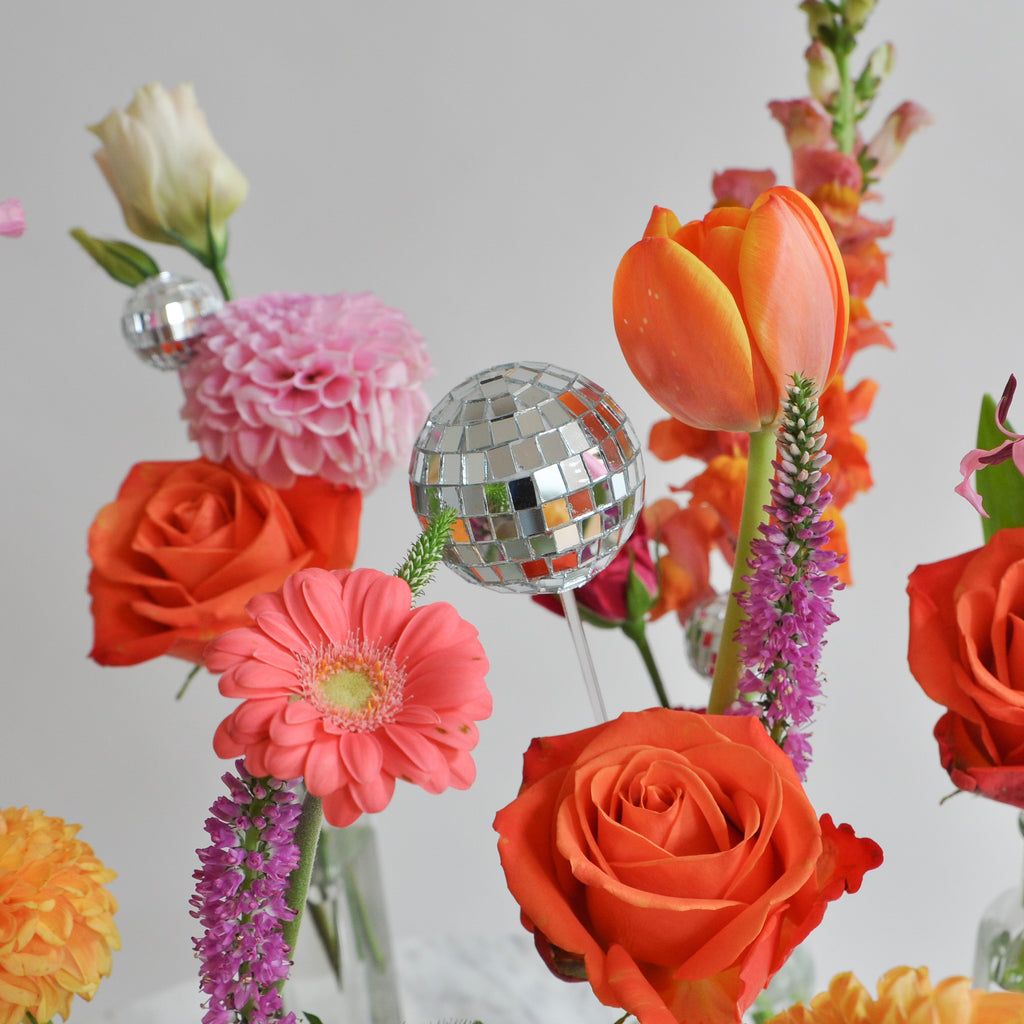 Close-up of a set of clear glass bud vases with tiny round, reflective mirror ball disco balls as well. The vases sit in front of a white backdrop on a marble table. They're filled with pink, orange, tangerine, and magenta-colored flowers, including rose, dahlia, piccolini daisy, tulip, and veronica and lily.