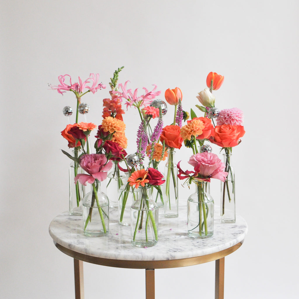 A set of clear glass bud vases with tiny round, reflective mirror ball disco balls as well. The vases sit in front of a white backdrop on a marble table. They're filled with pink, orange, tangerine, and magenta-colored flowers, including rose, dahlia, piccolini daisy, tulip, and veronica and lily.