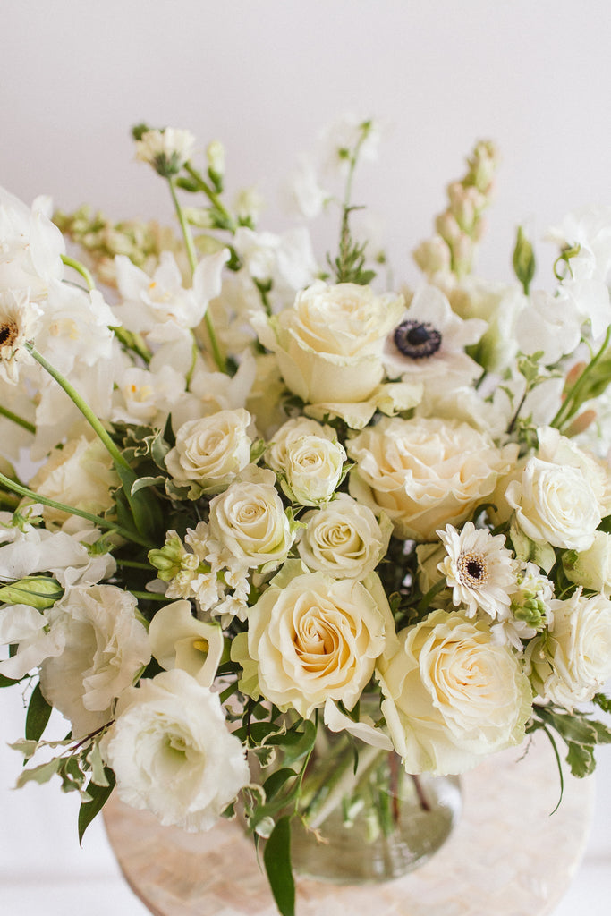 close-up of A flower arrangement in a glass container in front of a white backdrop on a small table. It's filled with white flowers, including rose, lisianthus, piccolini daisy, snapdragon, and anemone.