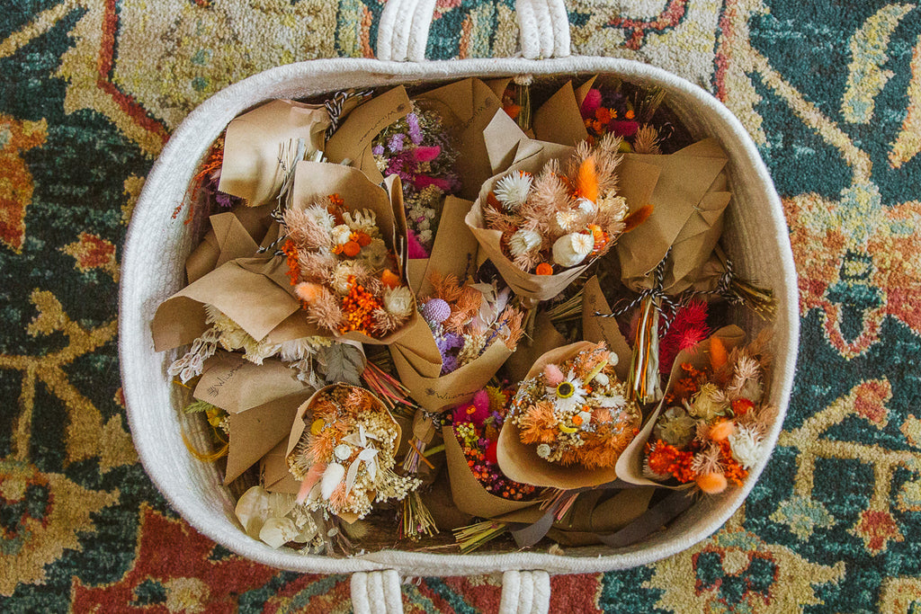 a basket full of mini dried floral bouquets about the size of a hand. they are in varying colors and wrapped in brown kraft paper that is stamped with a WildFlora logo. the basket sits on a teal tan and red rug.