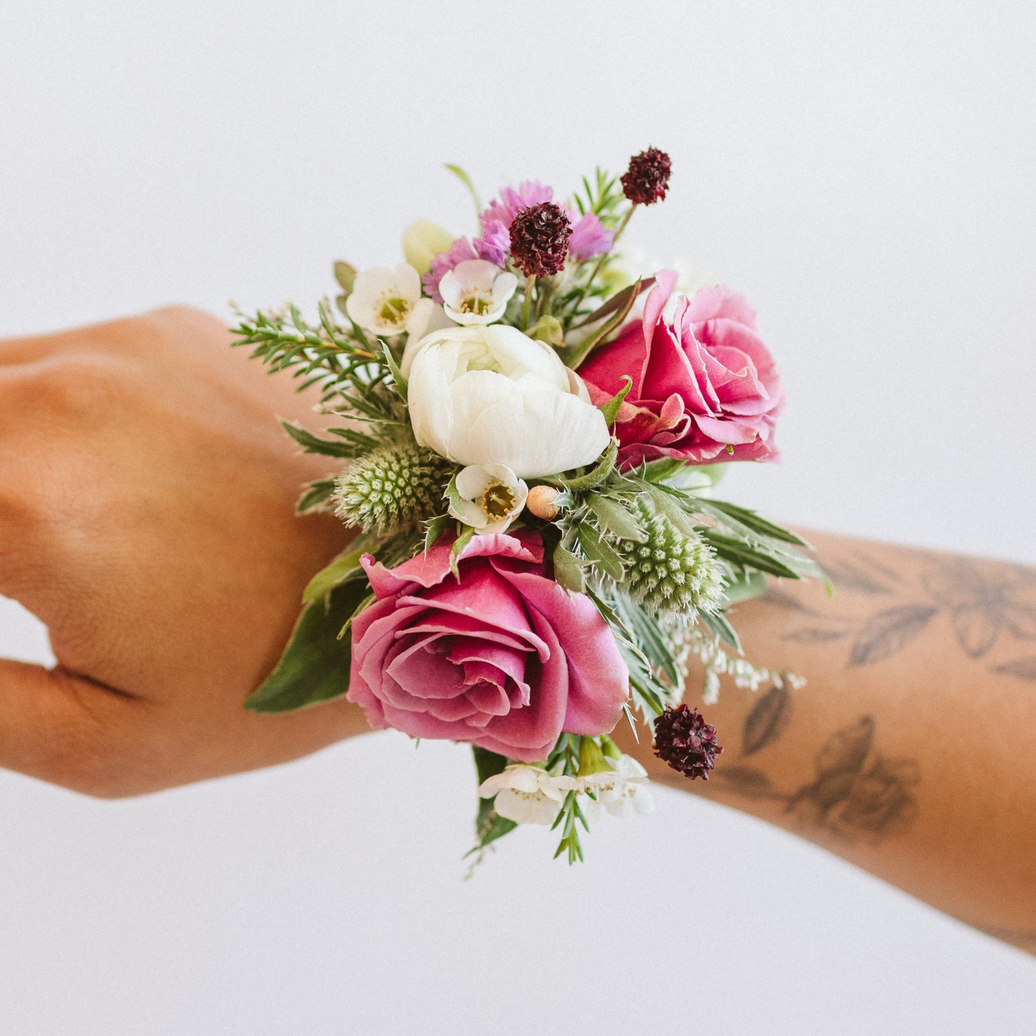 a tattooed arm wears a flower corsage in front of a white backdrop. It's made of pink, white, green, and dark purple flowers, including spray rose, thistle, ranunculus, and other foliage.