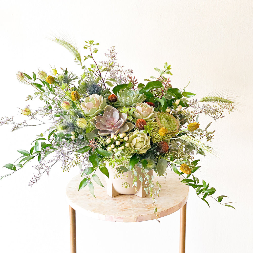 A flower arrangement in a white container in front of a white backdrop on a small table. It's filled with green and warm-colored flowers and foliage, including succulents, hypericum berries,, ornamental grass, thistle, and italian ruscus. It's got a wild, natural vibe to it.