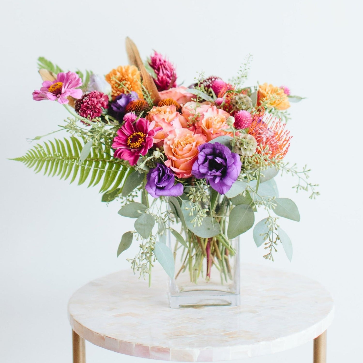 A glass vase with a small flower arrangement sits on a marble table in front of a white backdrop. The flowers are purple, magenta, orange, coral, and pink, with lush greens spilling around. The flowers included are lisianthus, pincushion protea, rose, scabiosa, and daisies.