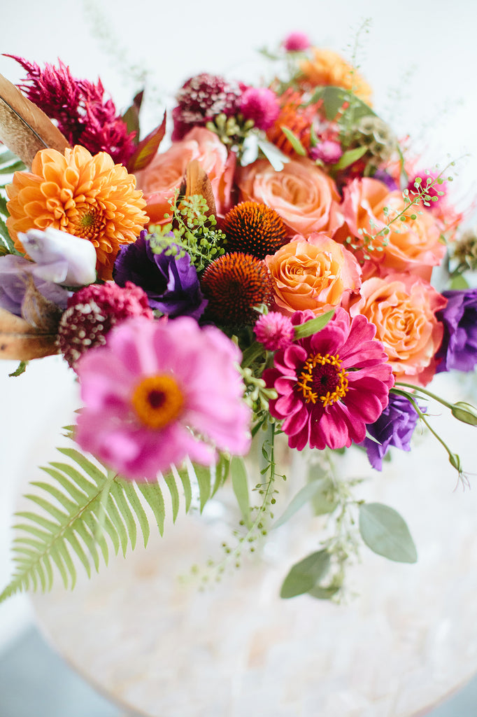 Close up of the blooms in a flower arrangement. The flowers are purple, magenta, orange, coral, and pink, with lush greens spilling around. The flowers included are lisianthus, pincushion protea, rose, scabiosa, and daisies.