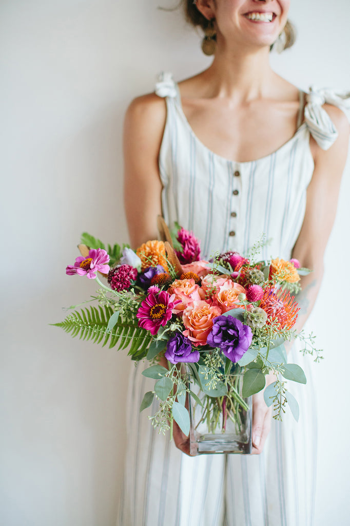 A woman in linen overalls holds a glass vase with a small flower arrangement in it. The flowers are purple, magenta, orange, coral, and pink, with lush greens spilling around. The flowers included are lisianthus, pincushion protea, rose, scabiosa, and daisies.