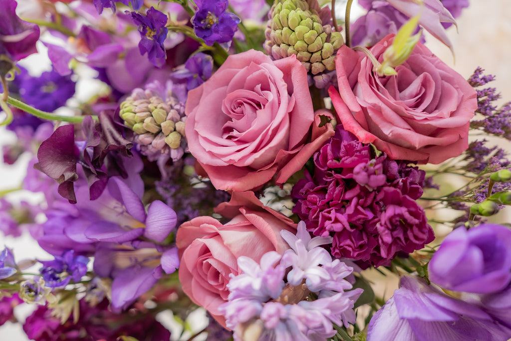 closeup of A flower arrangement in a glass container in front of a white backdrop on a small table. It's filled with lavender and purple flowers, including rose, stock, delphinium, and statice, hyacinth, and clematis.